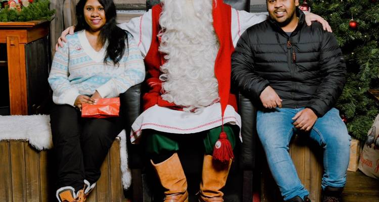 Two visitors pose with Santa Claus seated in a festive setting of Christmas trees and gifts.