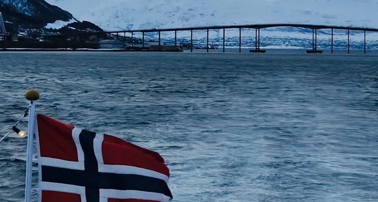 Norwegian flag flutters on a boat with long bridge and snow-covered mountains across cold waters.