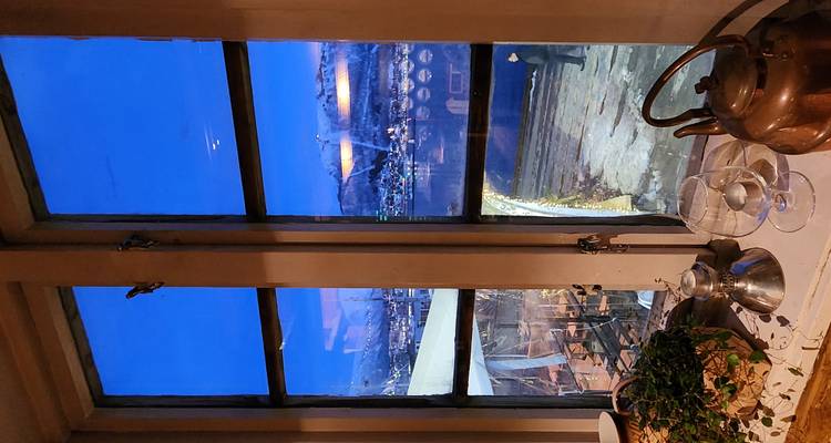 Evening harbour and mountain scenery seen through a cosy restaurant window with kettles and glasses in the foreground.