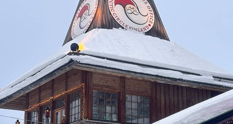 Wooden alpine building topped with the Santa Claus Village Arctic Circle Finland sign, roof blanketed in snow.
