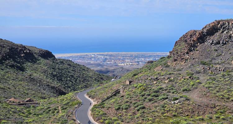 Route sinueuse à travers des pentes volcaniques verdoyantes s'ouvrant sur une ville côtière panoramique et la mer au-delà.