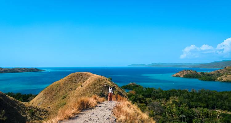 Traveler standing on a dusty ridge overlooking turquoise bays, small green islets and distant hills under a bright blue sky.