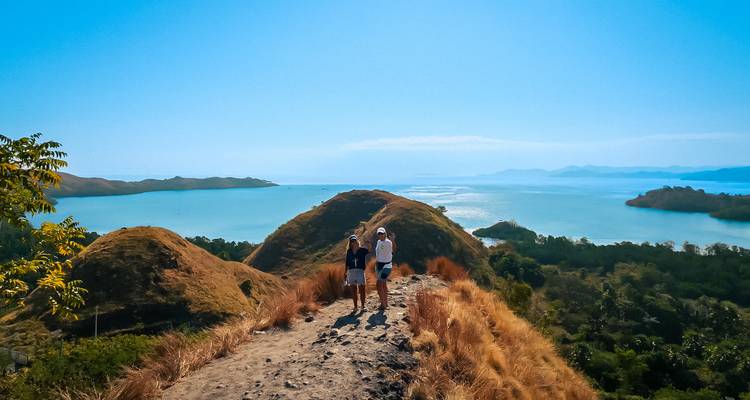 Two visitors walking along a narrow hilltop trail with sweeping views of blue sea and rolling islets.
