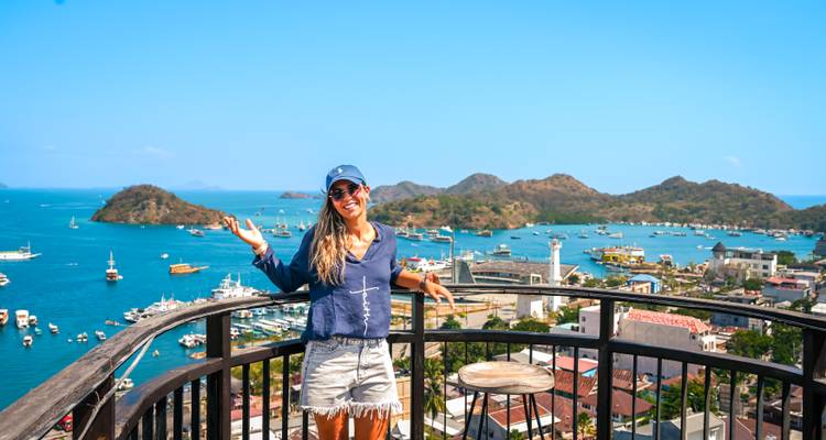 Smiling traveller on a viewpoint balcony overlooking Labuan Bajo harbour filled with boats and islands.