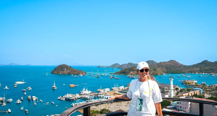 Smiling woman in white cap at a lookout railing with turquoise bay and green isles behind.