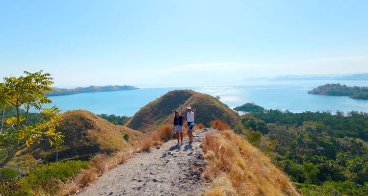 Two hikers walk along a dry grassy ridge overlooking bright blue island-dotted waters.