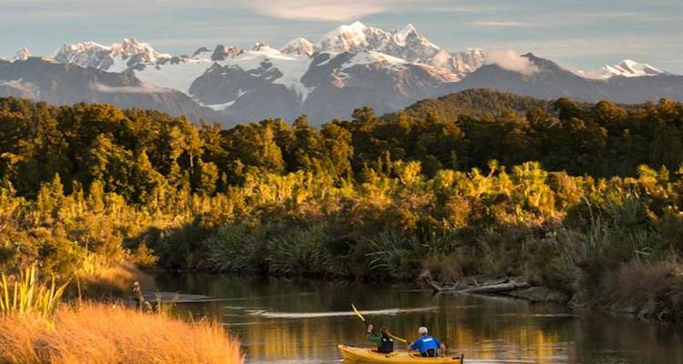 Deux personnes font du kayak sur une rivière calme avec des pics enneigés qui se dressent en arrière-plan.