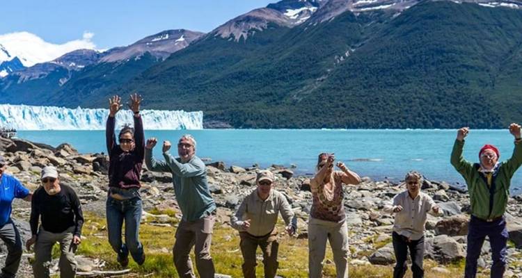 Les voyageurs bondissent avec joie sur la rive rocheuse d'un lac turquoise avec un glacier en arrière-plan.