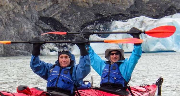 Deux kayakistes lèvent leurs pagaies triomphalement près d'un glacier dans des eaux grises et glaciales.