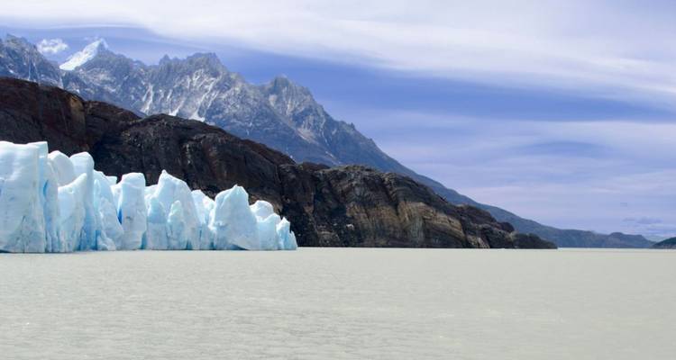 Enorme pared de glaciar azul encontrándose con un lago gris bajo los picos dentados de la Patagonia.
