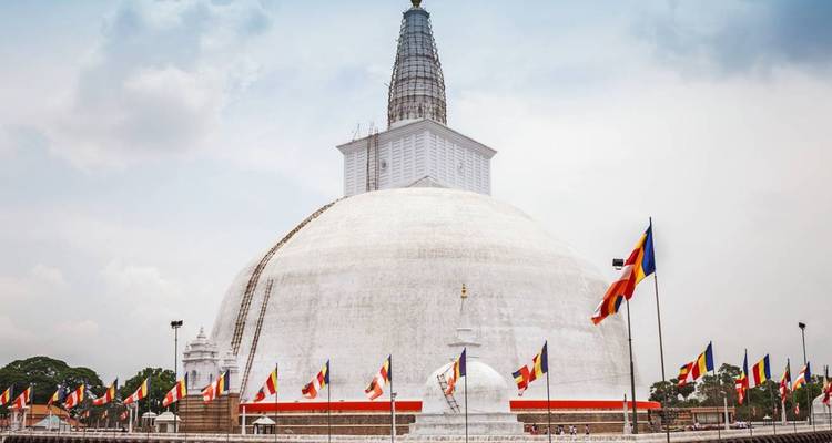 Massive white stupa Ruwanwelisaya under cloudy sky with colourful flags.
