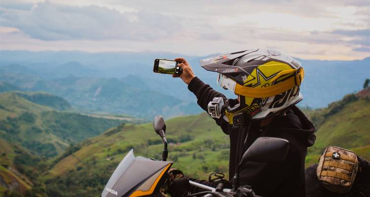 Ein Motorradfahrer mit Helm hält inne, um sich selbst vor einem Motorrad zu fotografieren, mit weitläufigen Bergpanoramen im Hintergrund.