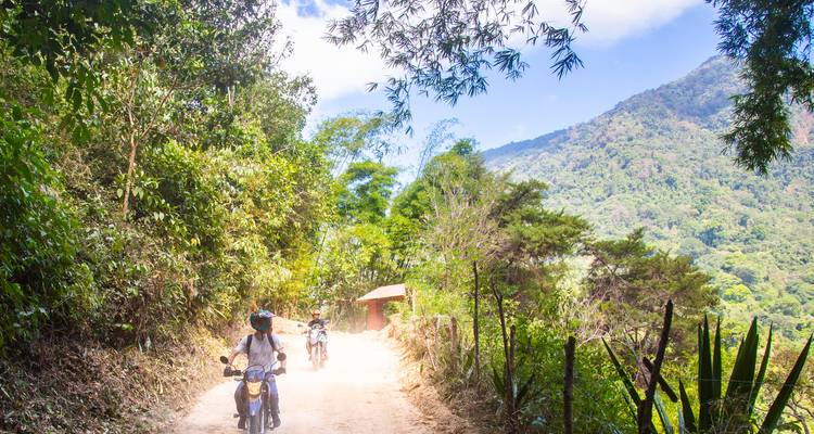 Three motorcyclists ride a dusty jungle path beneath a bright blue sky and forested hills.
