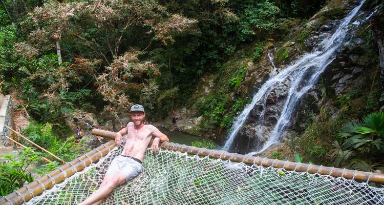 A man lounges shirtless on a rope net platform overlooking a cascading jungle waterfall.
