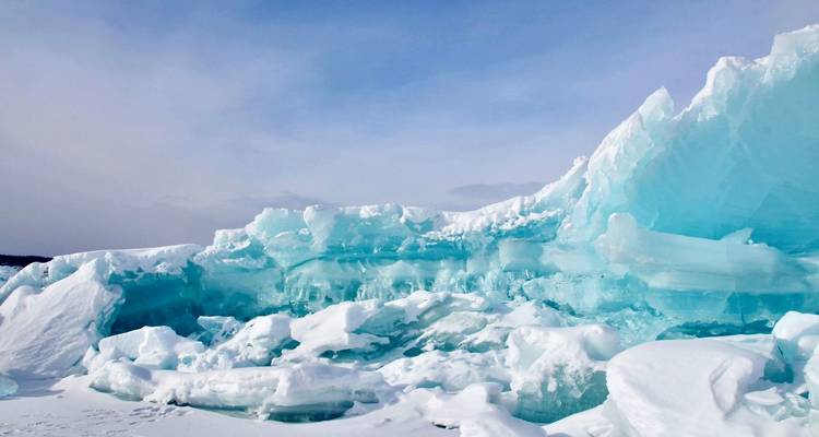 Vast wall of turquoise glacier ice with sculpted ridges rises from a snow-covered landscape under pale sky.