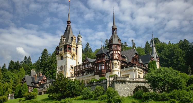 Château élégant de style néo-renaissance avec de hautes flèches situé sur un versant boisé sous un ciel partiellement nuageux.