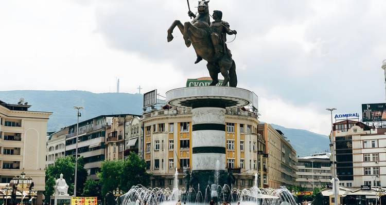 Grande statue équestre s'élevant au-dessus d'une fontaine dans une place publique animée, encadrée par des bâtiments environnants et des collines.