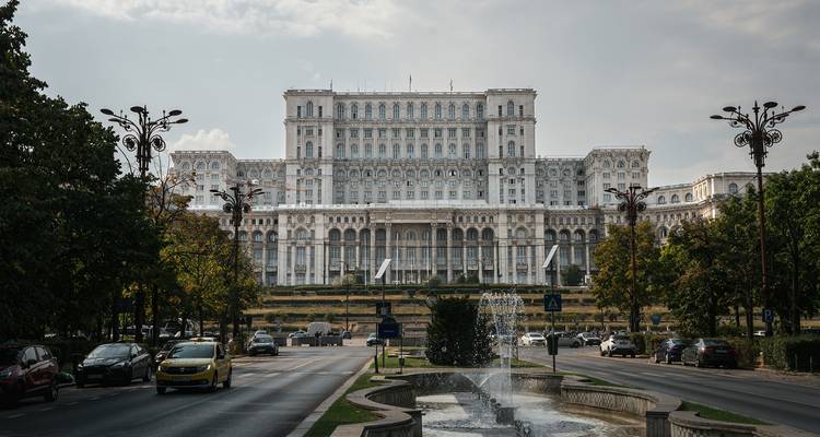 Un imposant bâtiment gouvernemental blanc domine une avenue urbaine bordée d'arbres et de fontaines.