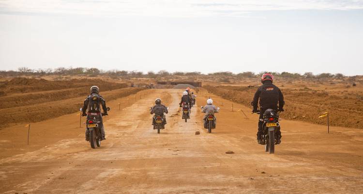 Un convoy de motociclistas baja por un ancho camino de tierra a través de un paisaje árido.
