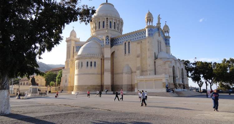 The white and blue domes of the Basilica of Our Lady of Africa stand against a clear sky while locals stroll nearby.