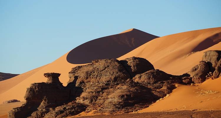 A towering sand dune casts a dramatic shadow over rugged desert rock formations.
