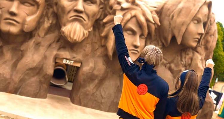 Two Naruto-clad fans raising fists toward the Hokage rock monument at a theme park.