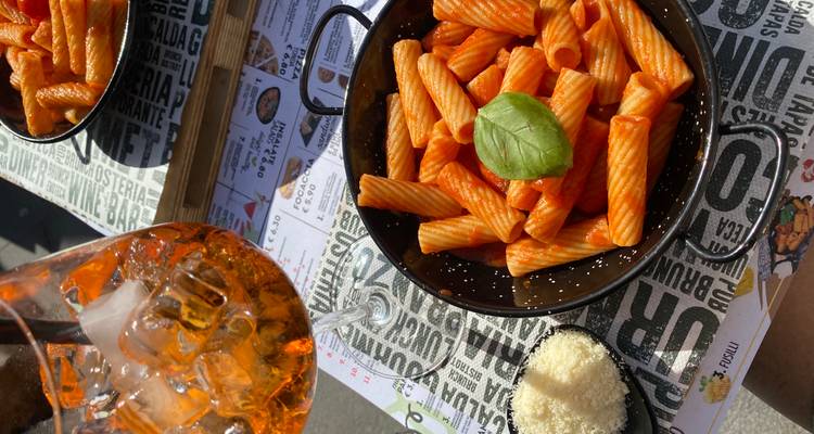 Kom rigatoni-pasta in tomatensaus met basilicum, geraspte kaas ernaast en een oranje spritz-cocktail op een tafel van een buitencafé