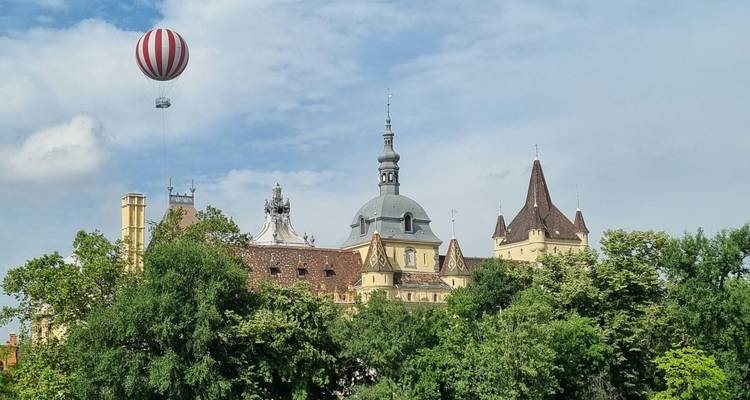 Historic castle with varied rooftops rising above green trees and a tethered hot-air balloon in the sky