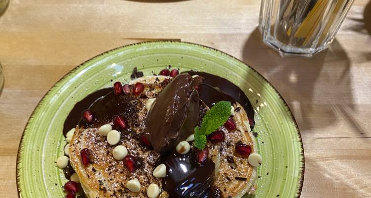 Plate of pancakes topped with chocolate sauce, pomegranate seeds, white chips and mint on wooden table
