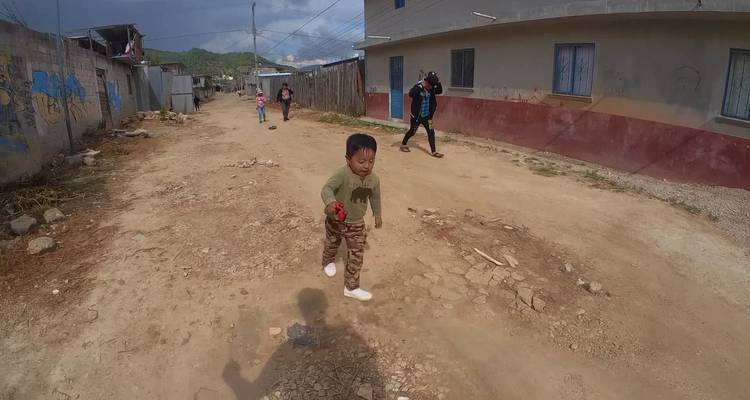 Children walk and play along a dusty unpaved street lined with modest village houses.