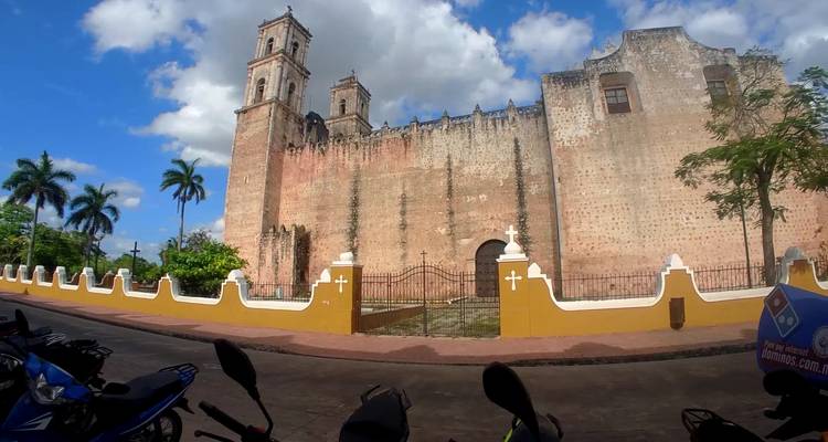 Wide-angle view of an old stone convent with twin bell towers set against a bright sky and palm trees.