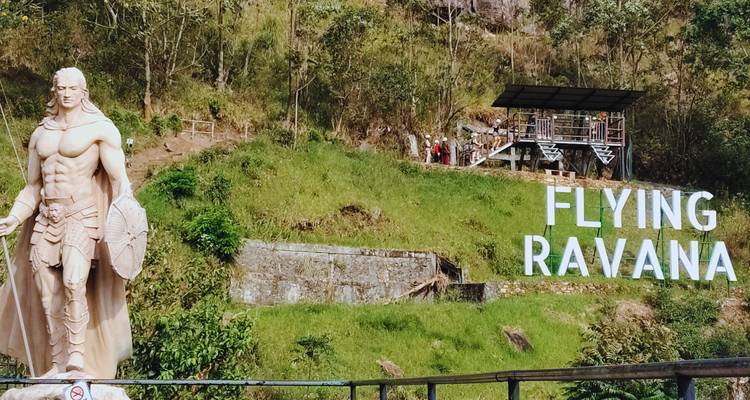 A hillside adventure park sign reads 'FLYING RAVANA' beside a heroic statue and green forested slopes.