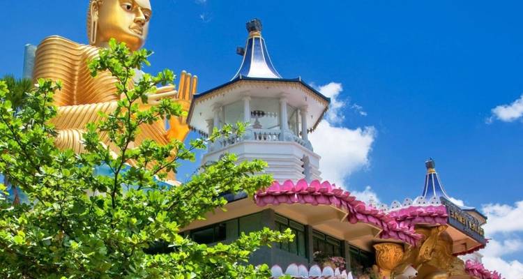 The massive golden Buddha statue towers above the colorful Dambulla Cave Temple complex under a vibrant blue sky.
