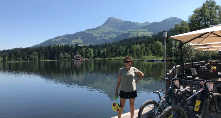 Une femme se tient sur un ponton au bord d'un lac avec des vélos de montagne tandis que les pics alpins et les forêts se reflètent sur un lac calme