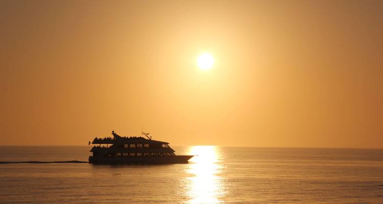 Small boat silhouette gliding across calm sea beneath a glowing orange sunrise.