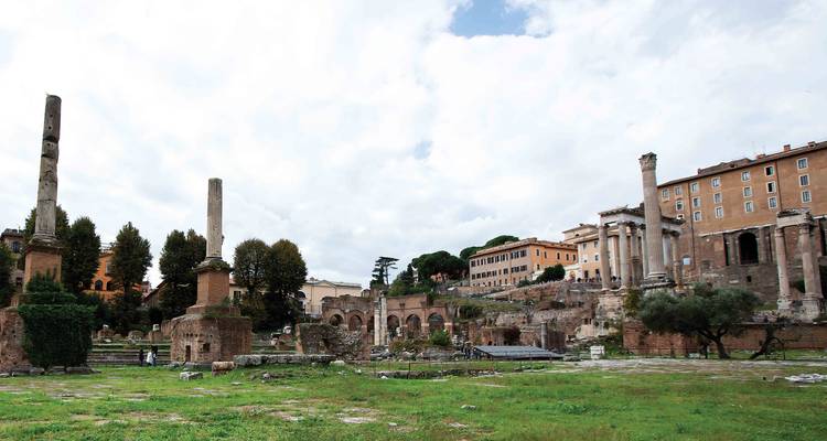Open grassy section of the Roman Forum with scattered columns and ruins under a bright cloudy sky.