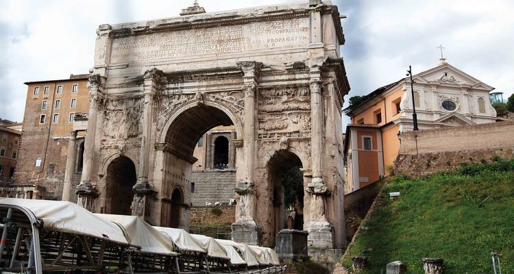 Arch of Septimius Severus stands majestically with weathered carvings beside grassy slopes and historic buildings.