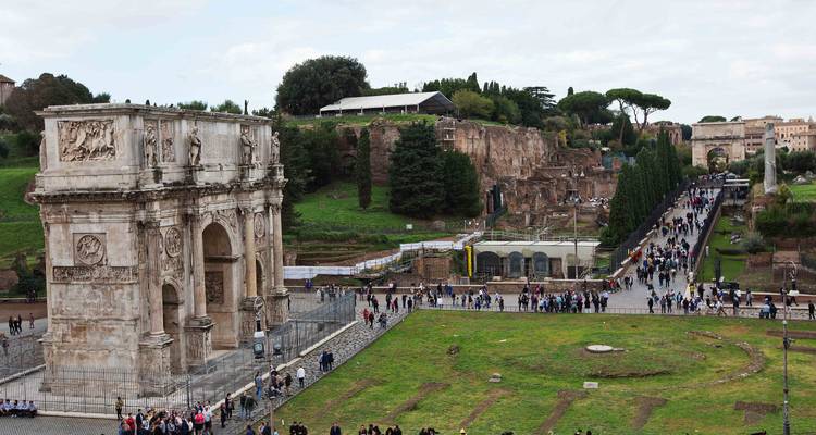 The Arch of Constantine stands beside pathways filled with visitors in the archaeological park near the Colosseum.