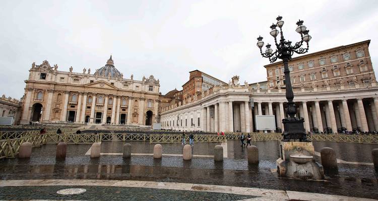 Wet cobblestones, colonnades and a vintage lamppost frame St. Peter’s Basilica under grey skies.