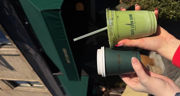 Mains tenant des boissons au café et au matcha à l'extérieur du Café Leon Dore sous un auvent vert.