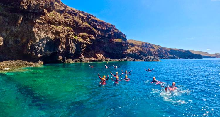 Large group in wetsuits snorkelling and swimming in vivid turquoise sea below rugged cliffs.