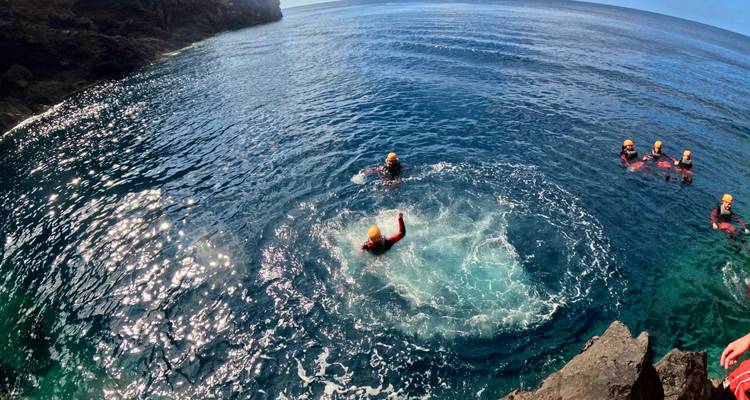 Participants in helmets and wetsuits floating and jumping into deep blue coastal water during coasteering.