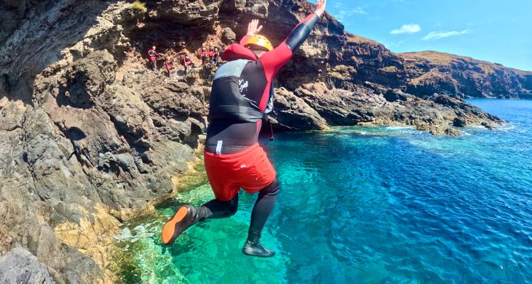 Adventurer in mid-air leap from rocky ledge into clear turquoise Atlantic cove.