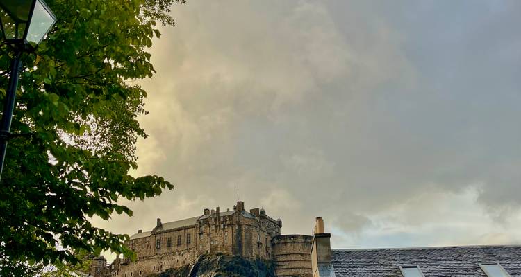 Le château d'Édimbourg illuminé par la lumière chaude du soir derrière les arbres feuillus et les toits.