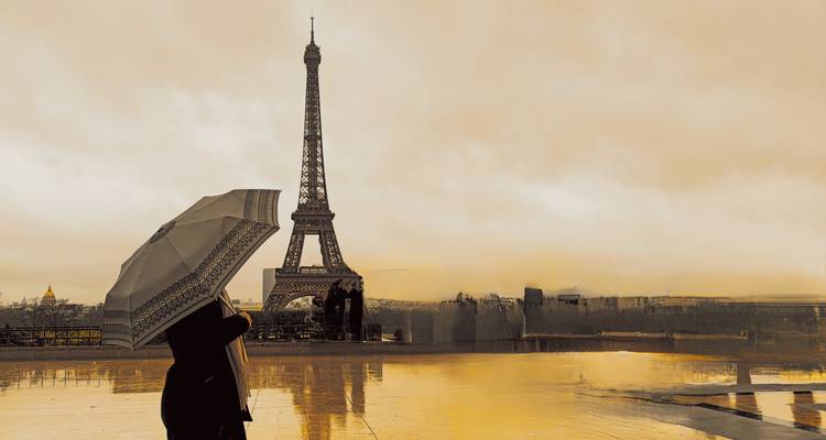 Silhouette of a person with an umbrella gazing at the Eiffel Tower reflected on a wet plaza in warm sunrise light.
