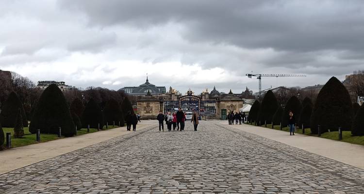 Long cobblestone avenue lined with conical hedges leading toward ornate golden gates under dramatic clouds.