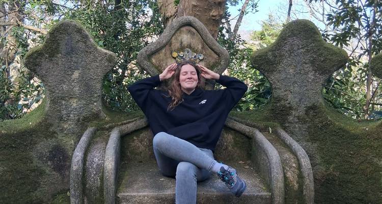 Young woman sitting playfully on moss-covered stone throne in forested garden.