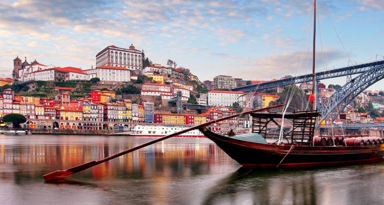 Un bateau rabelo traditionnel en bois flotte sur le fleuve Douro avec le bord de mer coloré de Porto et le pont Dom Luís I en arrière-plan.