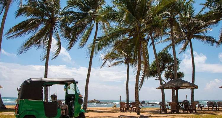A green tuk-tuk is parked on a sandy tropical beach lined with swaying palm trees beside the turquoise Indian Ocean.