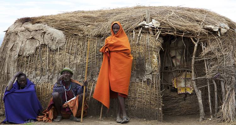 Trois villageois Maasai vêtus de shukas colorés se reposant à l'extérieur d'une hutte traditionnelle en terre.